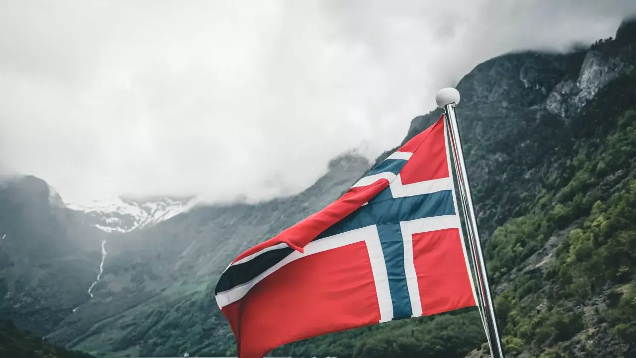Flaga Norwegii powiewa na tle mglistych gór i fiordu.