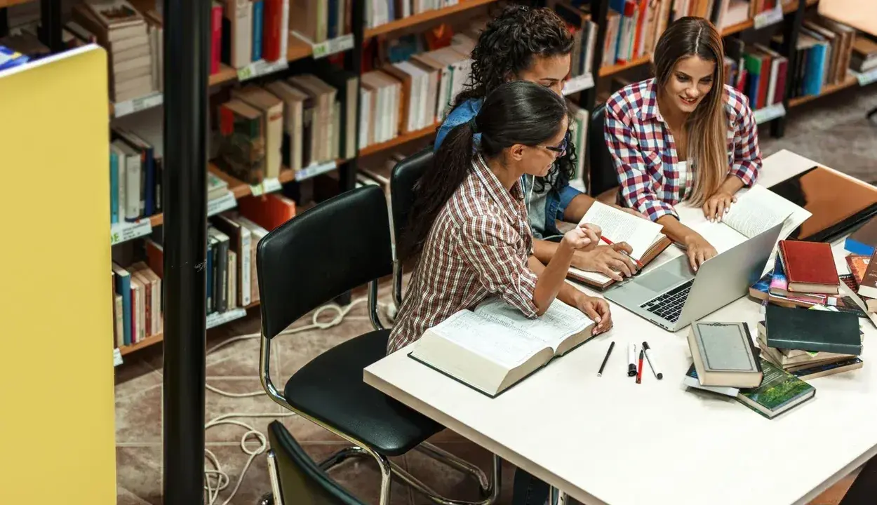 Trzy studentki przy stole w bibliotece, otoczone książkami, dyskutują o swoich kierunkach studiów.