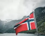 Flaga Norwegii powiewa na tle mglistych gór i fiordu.