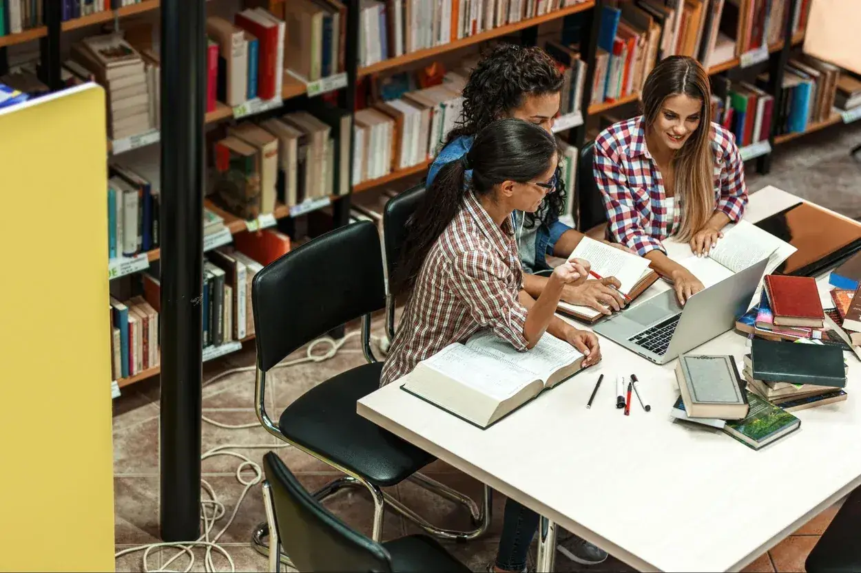 Trzy studentki przy stole w bibliotece, otoczone książkami, dyskutują o swoich kierunkach studiów.