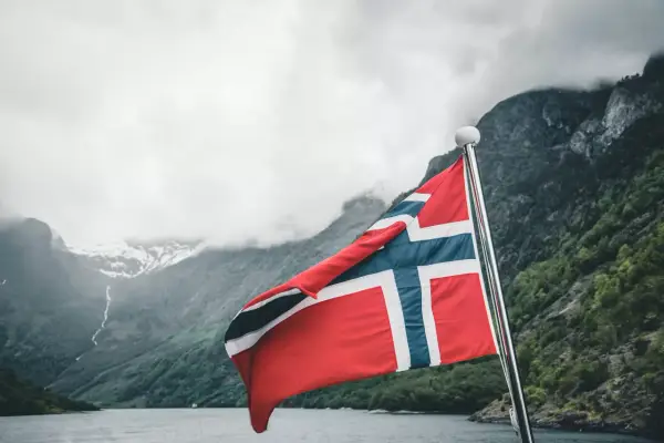 Flaga Norwegii powiewa na tle mglistych gór i fiordu.
