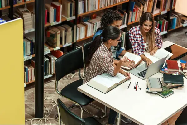 Trzy studentki przy stole w bibliotece, otoczone książkami, dyskutują o swoich kierunkach studiów.